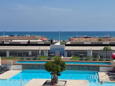Vue panoramique sur les piscines à plusieurs niveaux du complexe, avec transats, gazon artificiel et vues directes sur la mer Méditerranée et la côte lointaine.