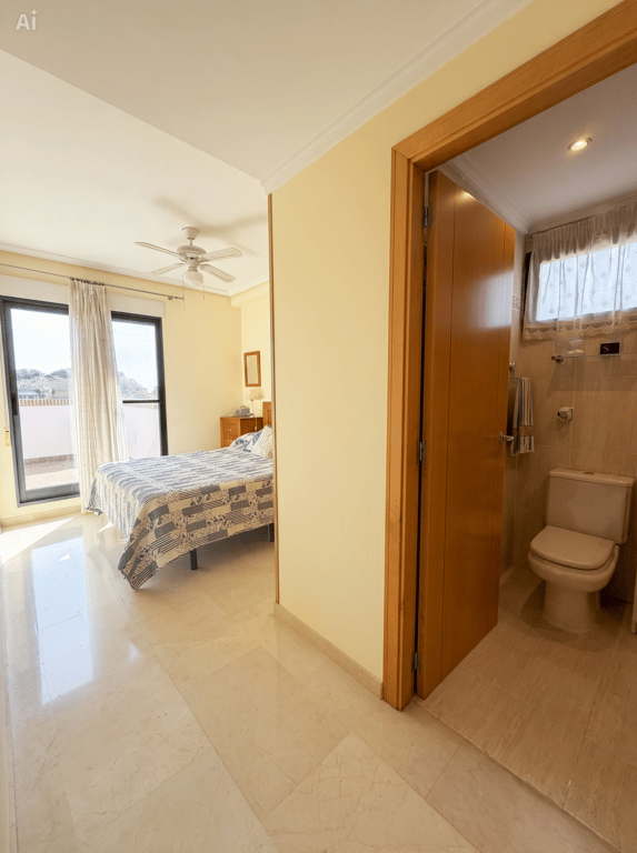 Chambre lumineuse avec sol effet marbre, lit double et ventilateur de plafond, s'ouvrant sur une terrasse par des portes vitrées. Une salle de bain carrelée avec toilettes est partiellement visible.