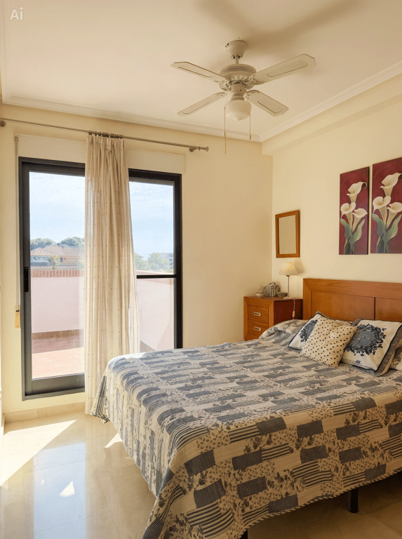Chambre lumineuse avec un lit double, une tête de lit en bois et un accès direct à une terrasse extérieure. Équipée d'un ventilateur de plafond et d'un sol carrelé clair.