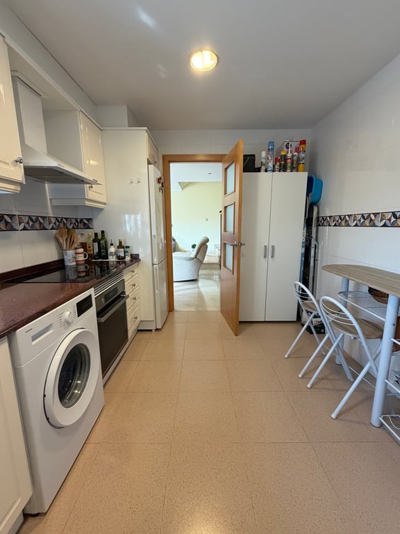 Cuisine fermée entièrement équipée avec armoires blanches brillantes, plan de travail foncé, lave-linge, four intégré et petite table à manger. Une porte en bois vitrée mène au salon adjacent.