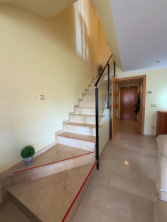 Escalier intérieur en marbre clair avec des bandes antidérapantes rouges et une balustrade moderne en métal noir et verre. Le sol carrelé assorti et la lumière naturelle complètent l'espace.