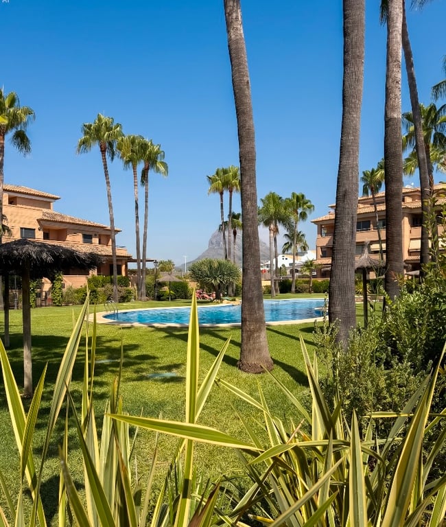La piscine communautaire est entourée de pelouses verdoyantes et de grands palmiers, offrant un oasis tropical avec parasols et bâtiments résidentiels sous un ciel bleu clair.