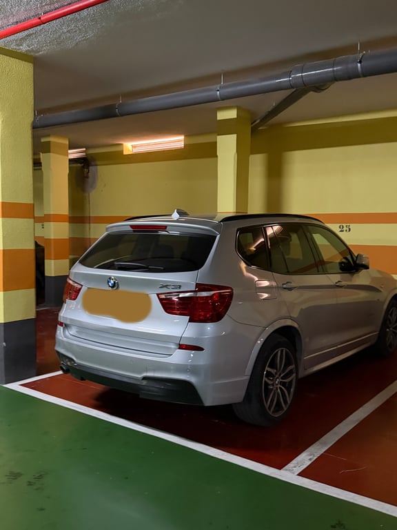Place de parking souterraine privée avec un SUV argenté, murs jaunes à rayures oranges et tuyaux rouges au plafond. Éclairage fluorescent et sol bicolore.
