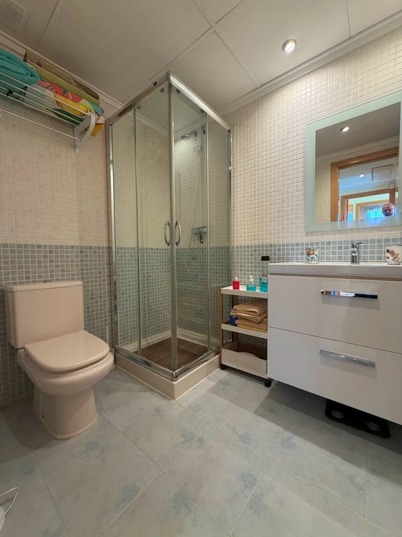 Salle de bain fonctionnelle avec cabine de douche vitrée, toilettes beiges et meuble-lavabo blanc avec miroir éclairé. Murs en mosaïque claire et bleue, assortie au carrelage du sol.