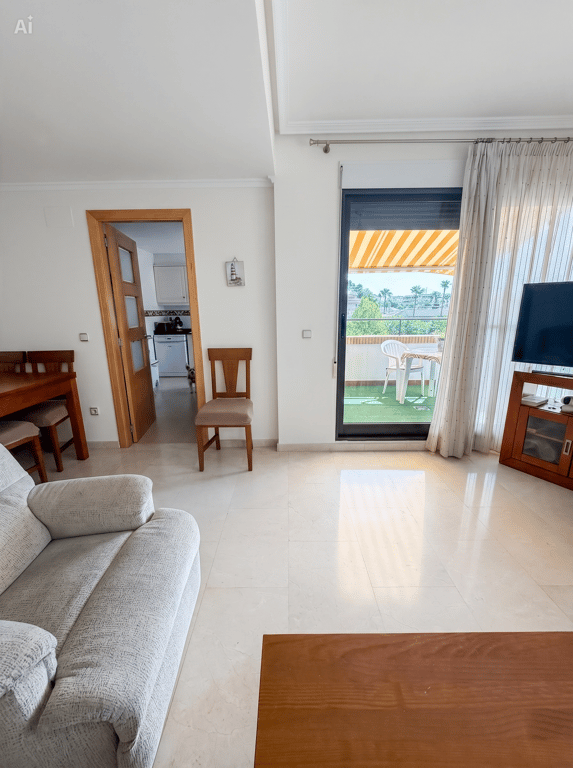 Séjour lumineux avec carrelage clair, un canapé et accès direct à une terrasse couverte avec gazon artificiel, mobilier extérieur et store rayé. Une porte mène à la cuisine.