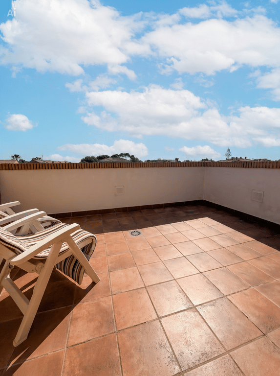 Vaste terrasse extérieure avec carrelage en terre cuite, un muret blanc surmonté de briques et deux chaises pliantes en bois, sous un ciel bleu nuageux.