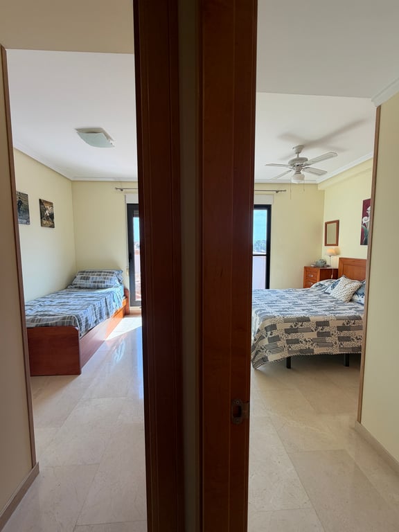 Vue depuis le couloir sur deux chambres avec carrelage en marbre clair et murs crème. La chambre principale dispose d'un lit double et d'un ventilateur de plafond, l'autre d'un lit simple.