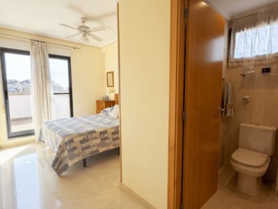 Chambre lumineuse avec sol effet marbre, lit double et ventilateur de plafond, s'ouvrant sur une terrasse par des portes vitrées. Une salle de bain carrelée avec toilettes est partiellement visible.