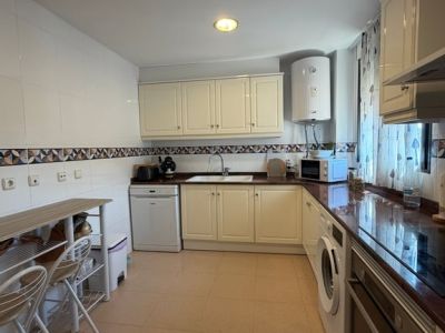 Cuisine fermée avec des armoires crème, des plans de travail foncés et des appareils électroménagers intégrés. Un petit bar en bois et des carrelages clairs sont présents.
