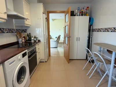 Cuisine fermée entièrement équipée avec armoires blanches brillantes, plan de travail foncé, lave-linge, four intégré et petite table à manger. Une porte en bois vitrée mène au salon adjacent.