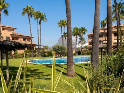 La piscine communautaire est entourée de pelouses verdoyantes et de grands palmiers, offrant un oasis tropical avec parasols et bâtiments résidentiels sous un ciel bleu clair.