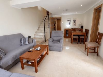 Le salon-salle à manger décloisonné offre un carrelage clair, un canapé gris confortable, une table basse en bois et un ensemble de salle à manger. Un escalier en marbre avec rampe noire mène à l'étage supérieur.