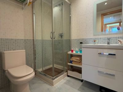 Salle de bain fonctionnelle avec cabine de douche vitrée, toilettes beiges et meuble-lavabo blanc avec miroir éclairé. Murs en mosaïque claire et bleue, assortie au carrelage du sol.
