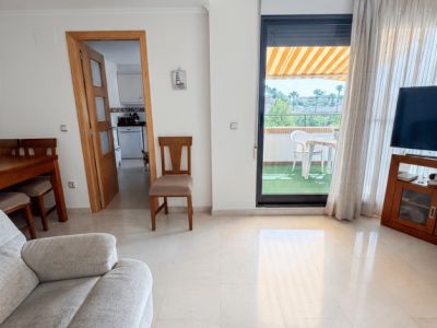 Séjour lumineux avec carrelage clair, un canapé et accès direct à une terrasse couverte avec gazon artificiel, mobilier extérieur et store rayé. Une porte mène à la cuisine.