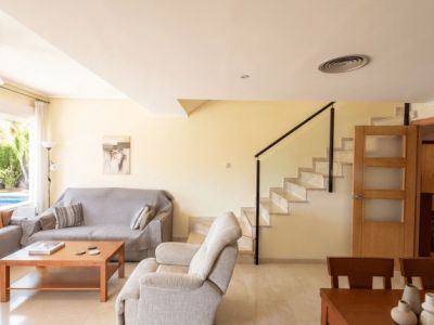Vaste espace salon-salle à manger doté d'un carrelage clair et d'une table en bois. Un escalier avec rampe noire mène à l'étage, et une fenêtre offre une vue partielle sur la piscine extérieure.