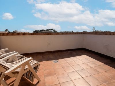 Vaste terrasse extérieure avec carrelage en terre cuite, un muret blanc surmonté de briques et deux chaises pliantes en bois, sous un ciel bleu nuageux.