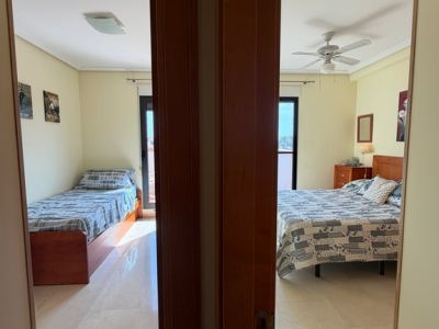 Vue depuis le couloir sur deux chambres avec carrelage en marbre clair et murs crème. La chambre principale dispose d'un lit double et d'un ventilateur de plafond, l'autre d'un lit simple.