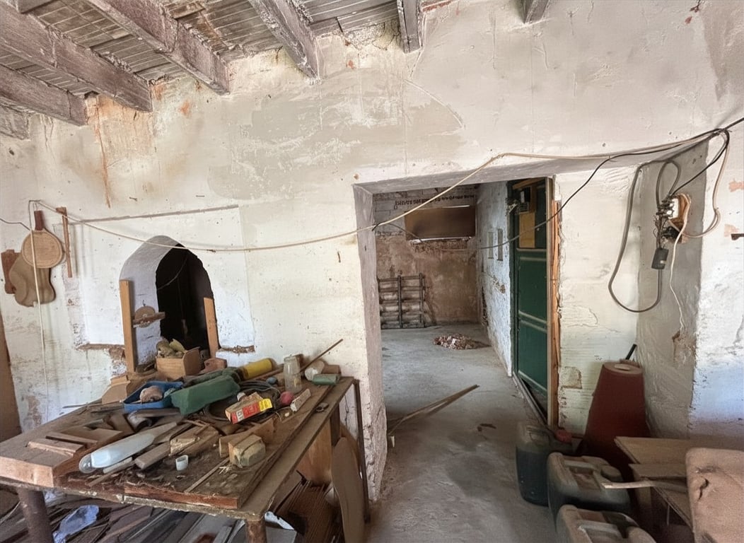 Intérieur à rénover avec poutres en bois apparentes et murs écaillés. Une alcôve cintrée est visible à gauche, devant un établi encombré d'outils divers. Intérieur à rénover avec poutres en bois apparentes et murs écaillés. Une alcôve cintrée est visible à gauche, devant un établi encombré d'outils divers.
