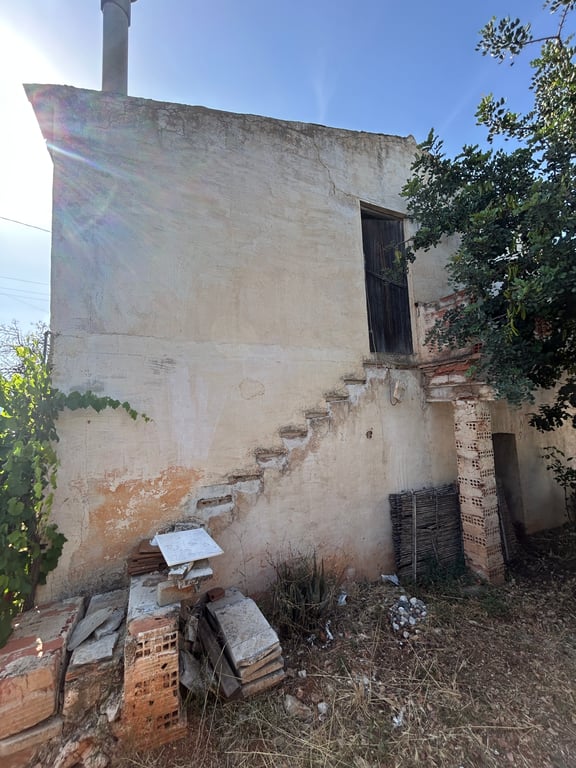 Vue extérieure de la propriété avec façade en stuc fissurée, escalier extérieur brisé menant à une porte en bois à l'étage, entouré de débris et de végétation clairsemée. Vue extérieure de la propriété avec façade en stuc fissurée, escalier extérieur brisé menant à une porte en bois à l'étage, entouré de débris et de végétation clairsemée.