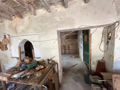 Intérieur à rénover avec poutres en bois apparentes et murs écaillés. Une alcôve cintrée est visible à gauche, devant un établi encombré d'outils divers.
