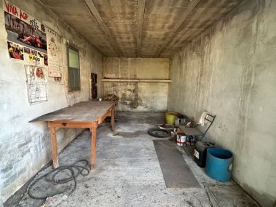 Intérieur d'un garage ou d'un entrepôt avec murs et plafond en béton brut. Une table en bois, une fenêtre et divers objets stockés révèlent un espace nécessitant une rénovation.