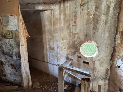 Intérieur présentant des murs vieillis avec briques apparentes, une ouverture ronde grillagée verte et une structure en bois brut. L'espace nécessite une rénovation complète.