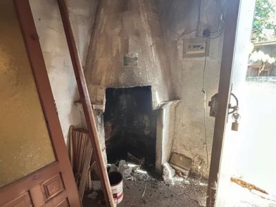 Pièce à rénover avec une cheminée maçonnée traditionnelle, murs bruts et débris au sol. Une porte en bois et une lumière naturelle entrent, révélant un potentiel de transformation.