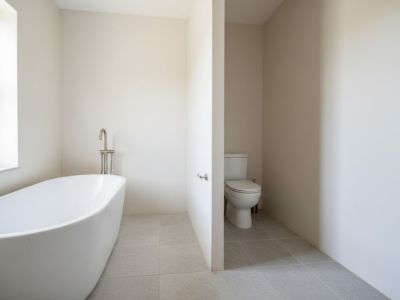 Salle de bain avec baignoire îlot blanche et robinet chromé sur pied. Le sol est carrelé en gris clair et une alcôve murale abrite les toilettes.