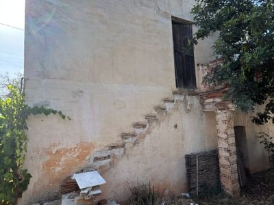Vue extérieure de la propriété avec façade en stuc fissurée, escalier extérieur brisé menant à une porte en bois à l'étage, entouré de débris et de végétation clairsemée.