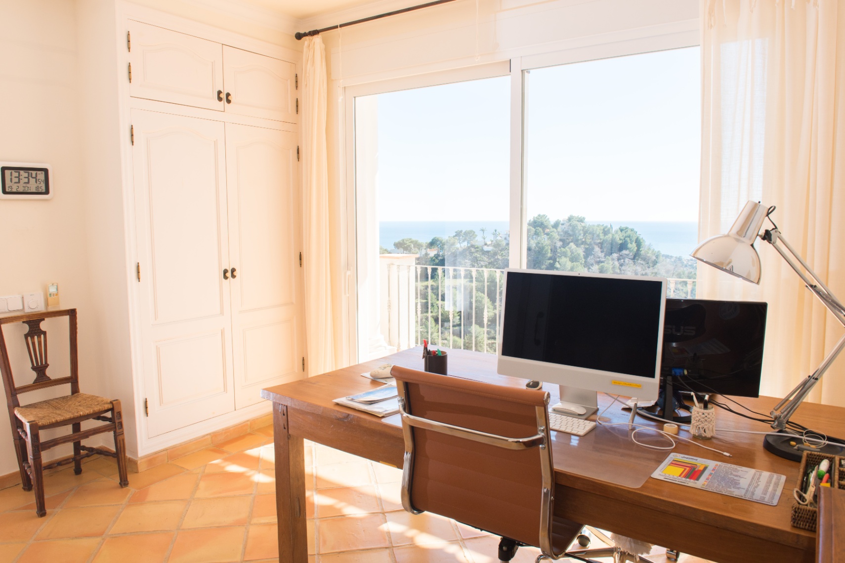 Bureau lumineux avec bureau en bois, armoires encastrées blanches et grandes baies vitrées offrant une vue panoramique sur la mer et la côte.
