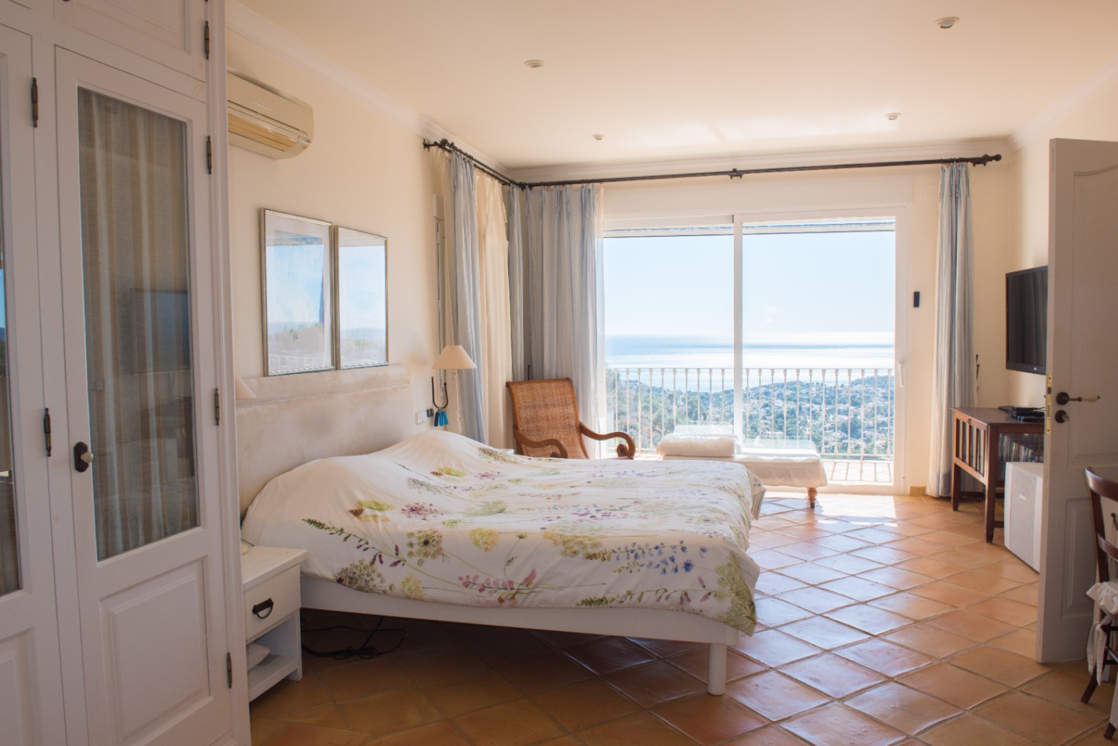 Chambre lumineuse avec sol en terre cuite, armoires encastrées blanches et accès direct à un balcon offrant une vue panoramique sur la mer Méditerranée.
