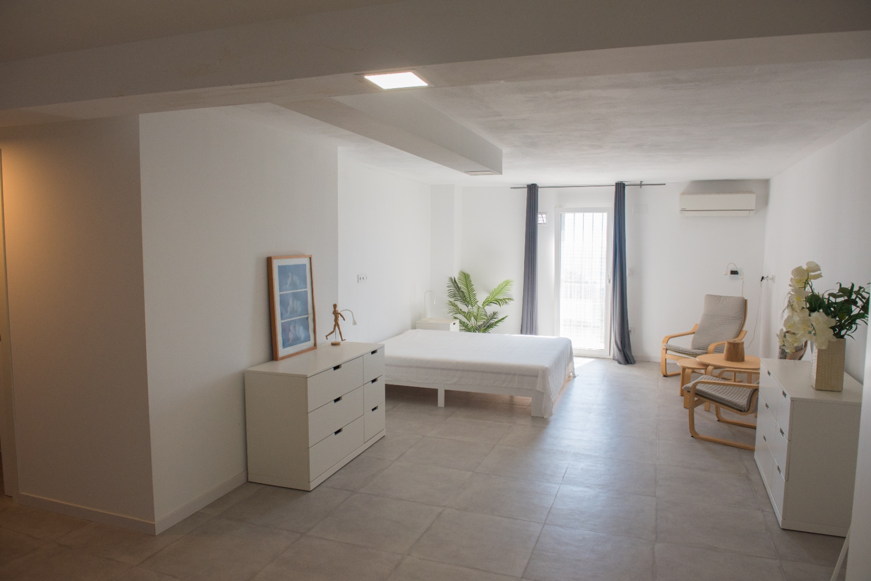 Chambre minimaliste aux tons neutres avec sol en céramique grise, mobilier blanc fonctionnel, climatisation et accès extérieur direct.