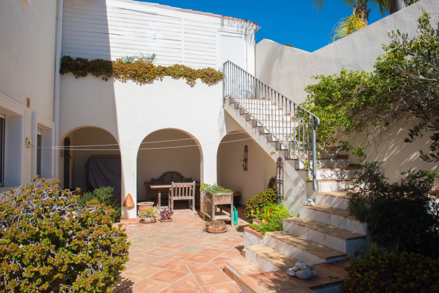 Cour extérieure avec sol en terre cuite, comprenant des alcôves voûtées ombragées et un escalier extérieur menant au niveau supérieur de la villa.
