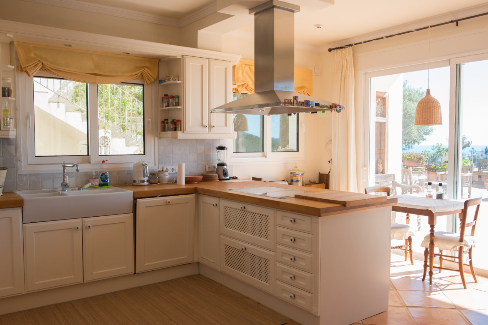 Cuisine de style rustique avec placards blancs, plans de travail en bois massif, évier en céramique et hotte en inox offrant un accès direct à la terrasse.