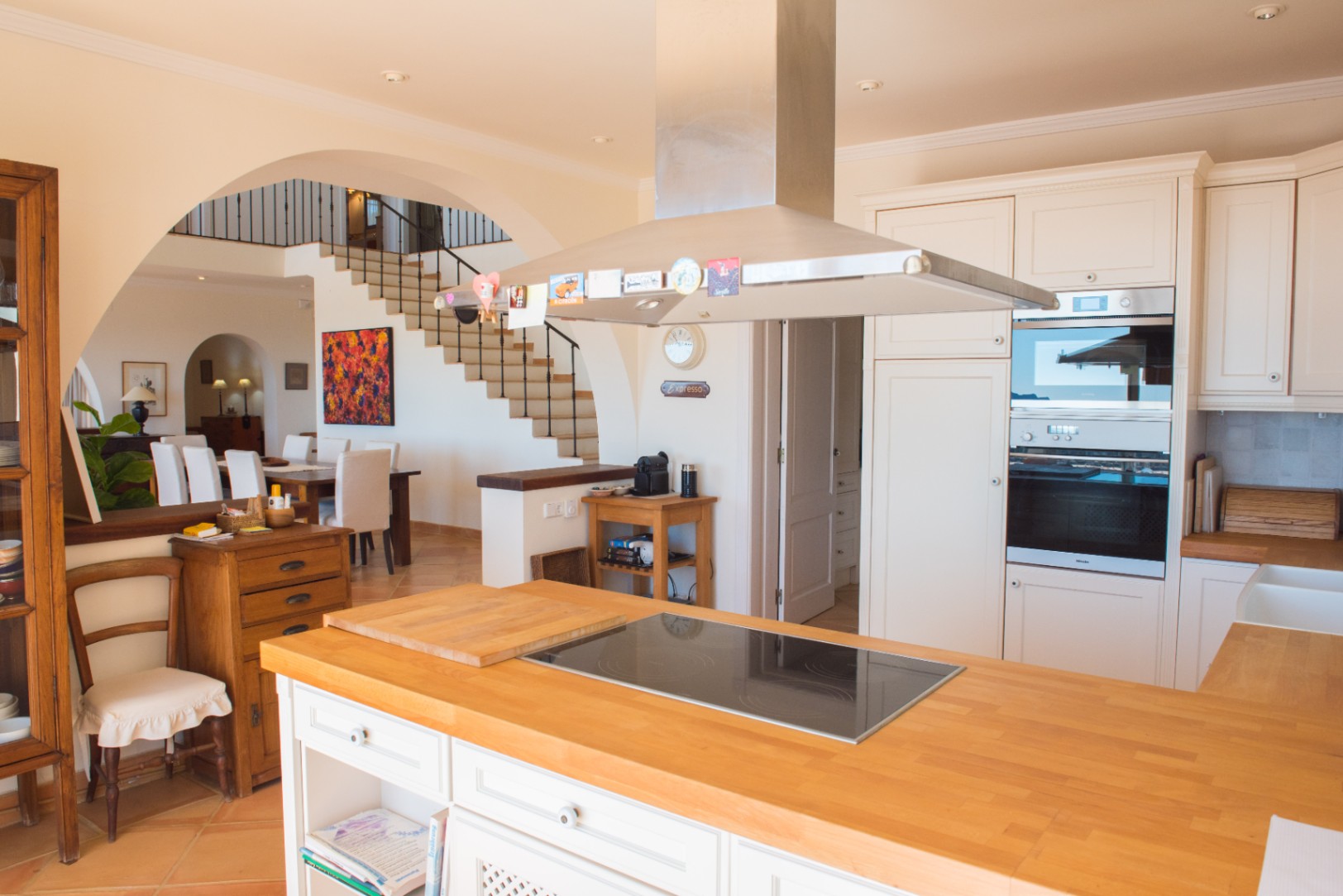 Cuisine ouverte avec îlot en bois, plaque à induction, hotte en inox et fours encastrés, offrant une vue sur la salle à manger et l'escalier via une arche.