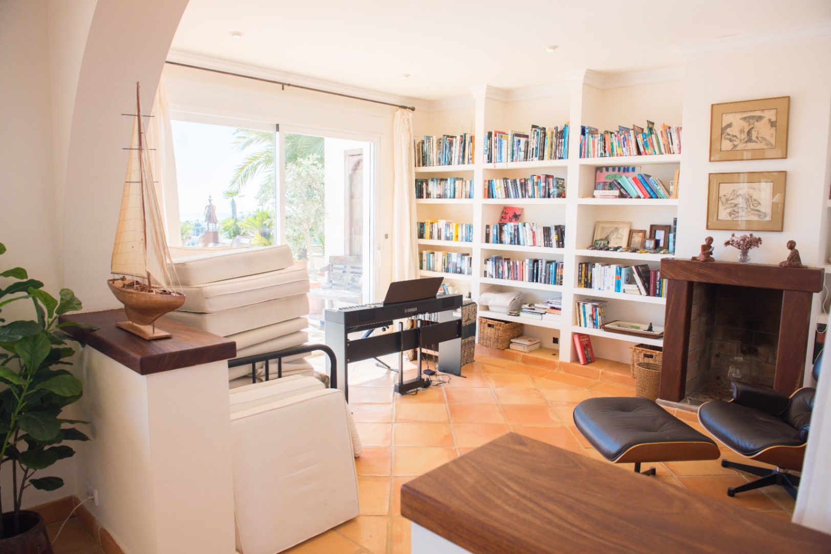 Espace bureau avec bibliothèque intégrée, sol en terre cuite, cheminée avec manteau en bois et accès direct à la terrasse ensoleillée.