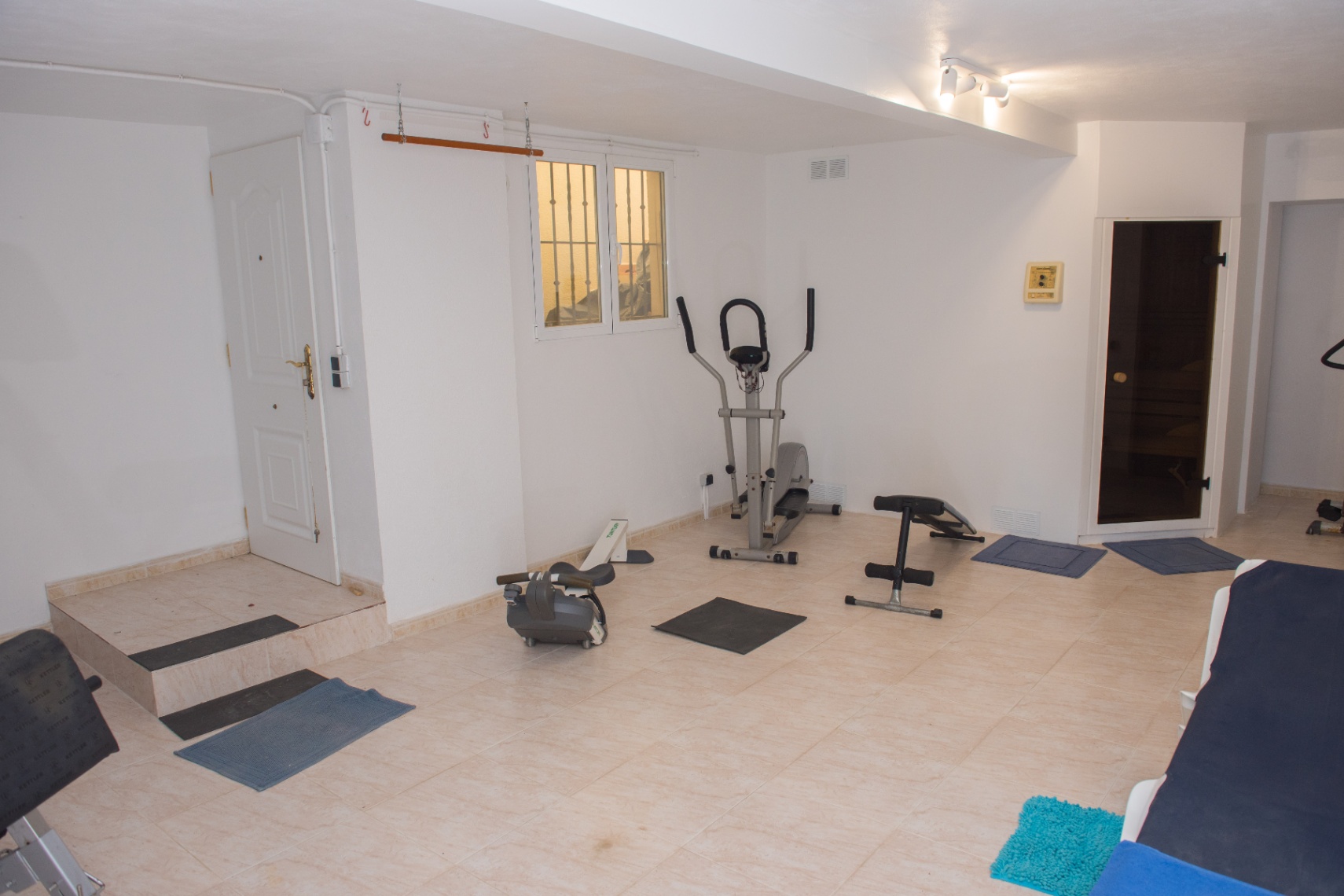 Espace fitness privé avec appareils de cardio, banc de musculation et accès direct à un sauna. La pièce dispose d'un sol en carrelage clair.