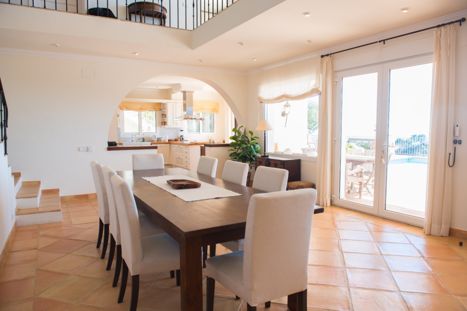 Salle à manger spacieuse avec table en bois massif, sol en terre cuite et arche menant à la cuisine, offrant un accès direct à la terrasse et à la piscine.
