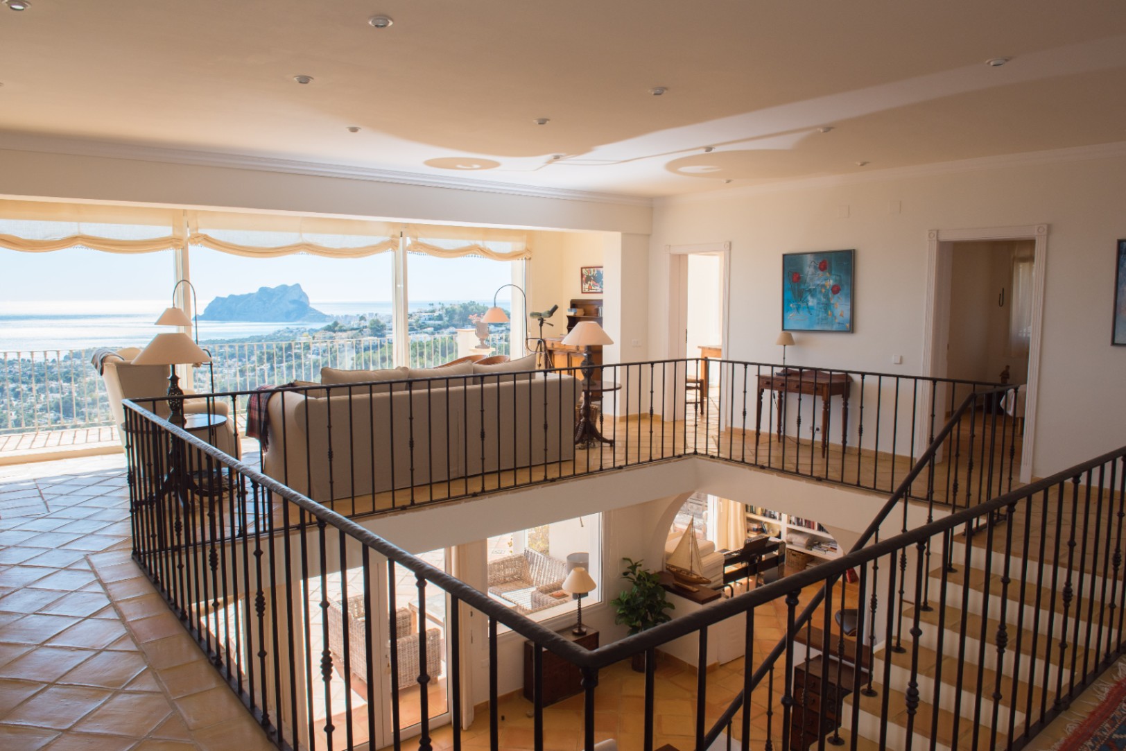 Salon en mezzanine avec garde-corps en fer forgé, carrelage en terre cuite et grandes baies vitrées offrant une vue panoramique sur la mer et le Peñón de Ifach.