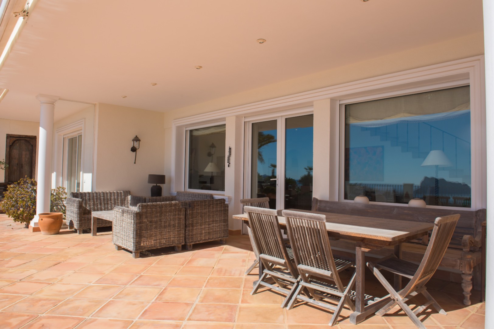 Terrasse couverte avec sol en terre cuite, espace repas en bois et salon en rotin, bordée de grandes baies vitrées.