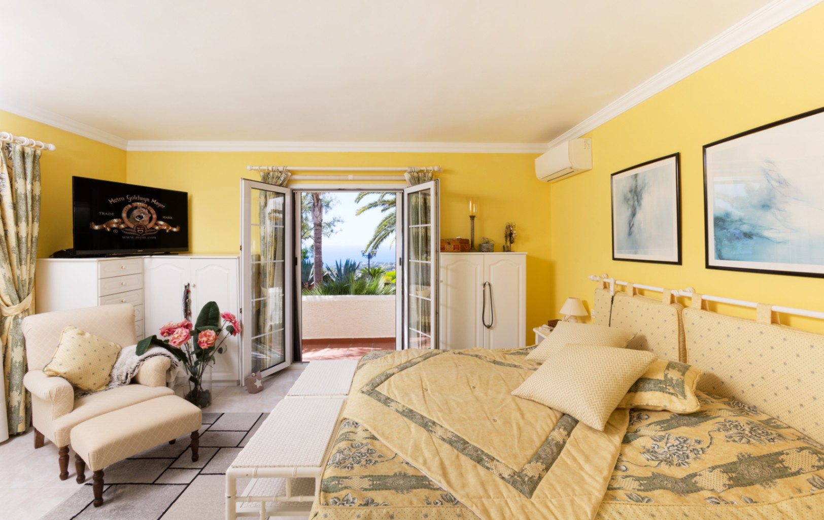 Chambre aux murs jaunes avec mobilier blanc et coin salon. Des portes vitrées s'ouvrent sur une terrasse privée offrant une vue sur la mer et des palmiers.