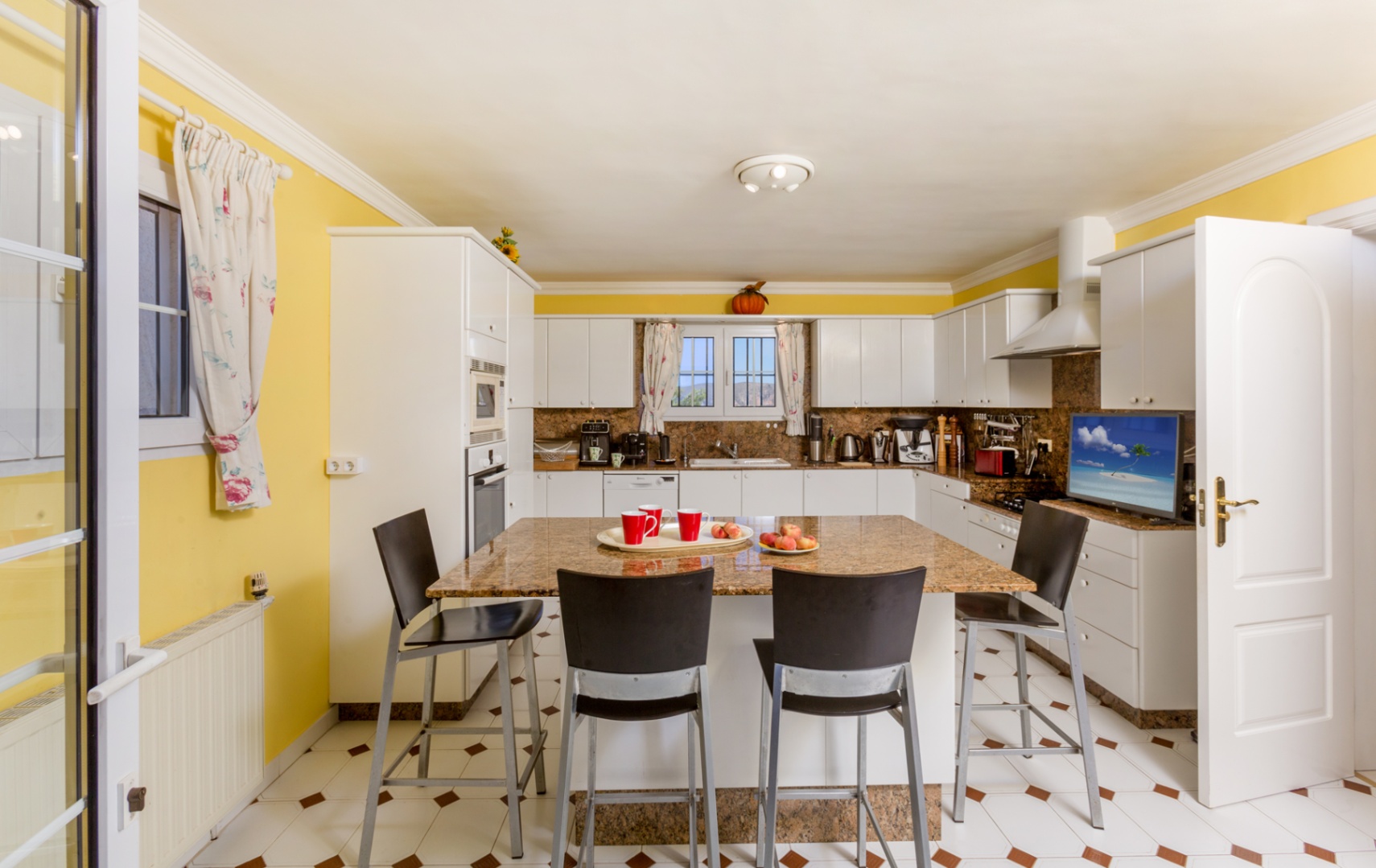 Cuisine spacieuse avec îlot central en granit, tabourets noirs, meubles blancs et murs jaunes. Le sol carrelé affiche des motifs décoratifs bruns.