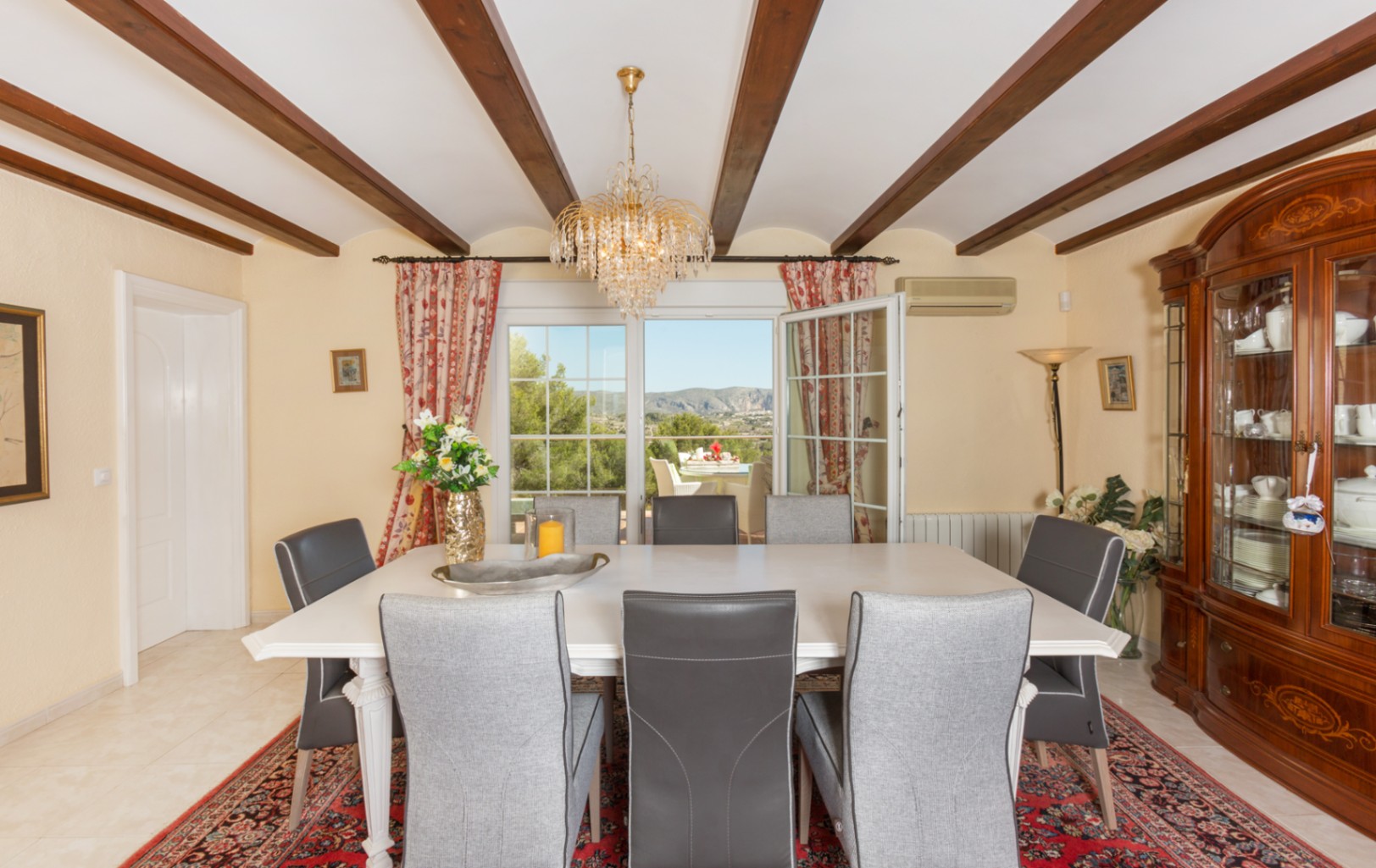 Salle à manger dotée de poutres apparentes, d'une table blanche et d'un buffet classique. Les portes vitrées offrent un accès direct à la terrasse et aux montagnes.