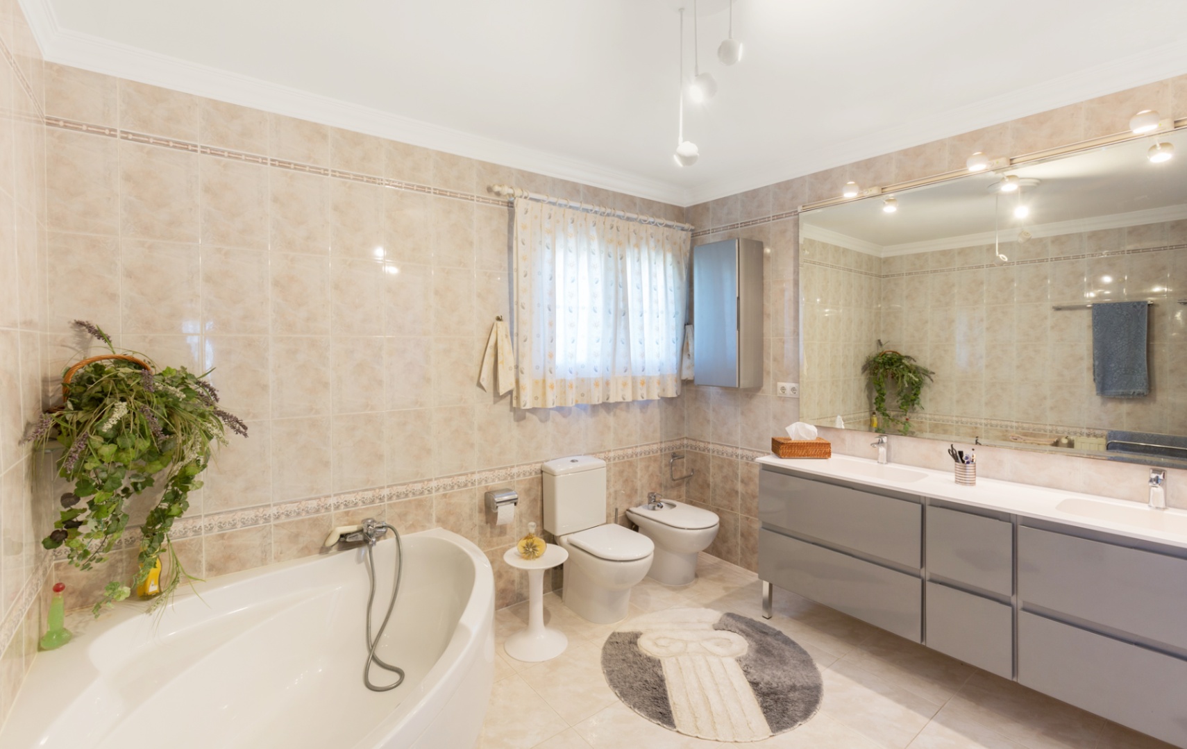 Salle de bain spacieuse avec baignoire d'angle, double vasque sur meuble gris et grand miroir. Murs et sols en carrelage beige avec frises décoratives.