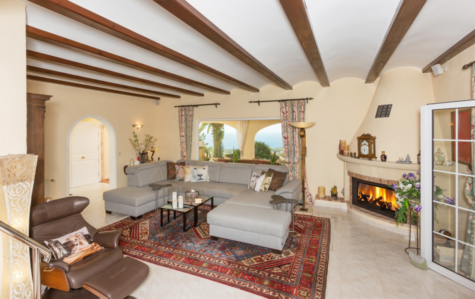 Salon spacieux avec poutres apparentes en bois, grand canapé d'angle gris, cheminée traditionnelle et vue sur la mer à travers la fenêtre.