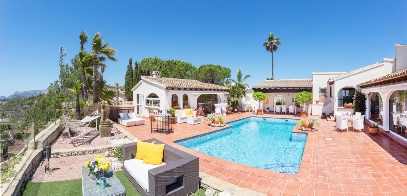 Terrasse avec piscine rectangulaire, revêtement en terre cuite et plusieurs coins salon. L'architecture méditerranéenne présente des murs blancs et des fenêtres cintrées.