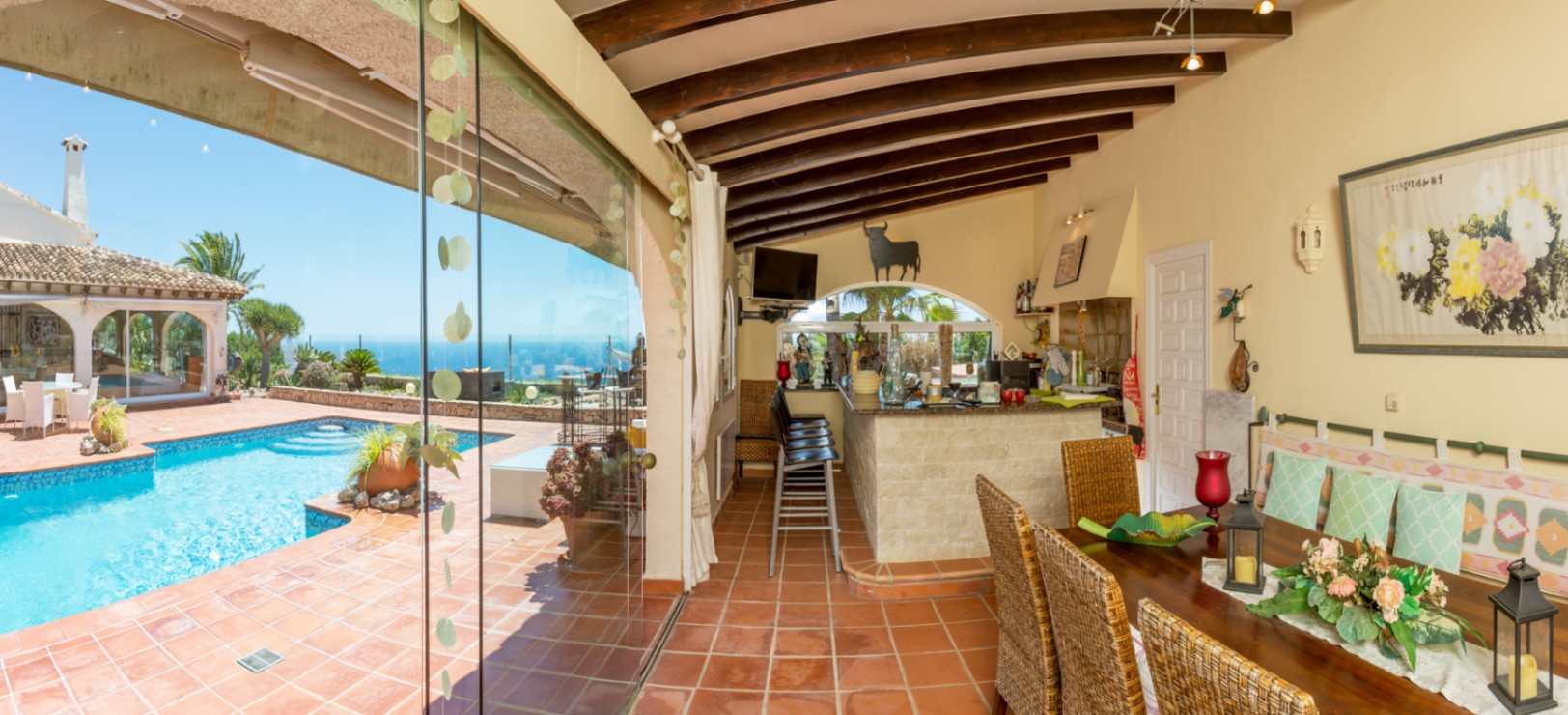 Terrasse couverte avec poutres apparentes, sol en terre cuite et bar extérieur en pierre, offrant une vue directe sur la piscine et l'horizon marin.