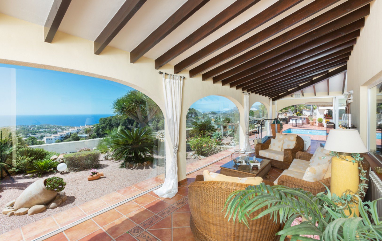 Terrasse couverte avec poutres en bois sombre, sol en terre cuite et salon en osier. Les arches vitrées s'ouvrent sur une vue panoramique sur la mer et le jardin.