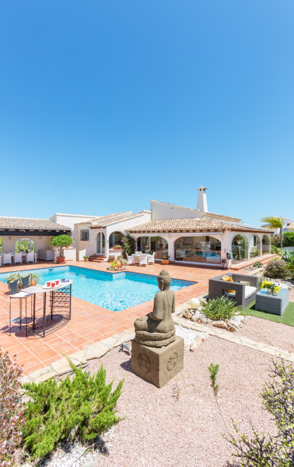 Terrasse extérieure avec piscine, revêtement en terre cuite, statue de Bouddha en pierre et villa méditerranéenne avec une véranda vitrée.