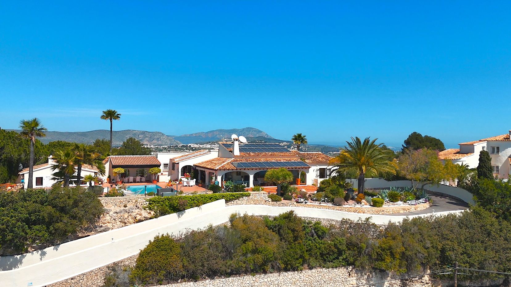 Vue aérienne de la villa avec toits en terre cuite, panneaux solaires et piscine, entourée de jardins méditerranéens face à la mer.